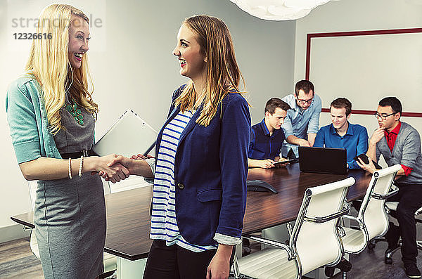 Eine junge Geschäftsfrau lobt ihre Mitarbeiterin für ihre Bemühungen  während ihre Kollegen ihre Arbeit in einem Konferenzraum an ihrem Firmensitz fortsetzen; Sherwood Park  Alberta  Kanada