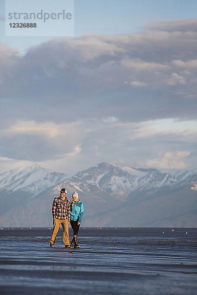 Ein junges Paar  das an einem Strand spazieren geht und sich an den Händen hält  mit einer Bergkette in der Ferne; Anchorage  Alaska  Vereinigte Staaten von Amerika