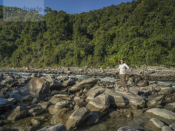 Eine Frau steht auf einem großen Felsen am Ufer eines Flusses mit einem üppigen Wald im Hintergrund; Westbengalen  Indien