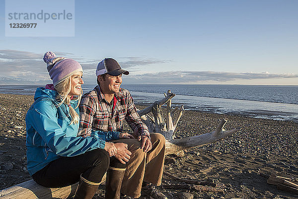 Ein junges Paar sitzt auf einem Stück Treibholz am Strand und blickt bei Sonnenuntergang auf den Ozean; Anchorage  Alaska  Vereinigte Staaten von Amerika