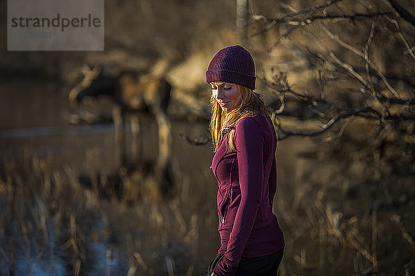 Eine junge Frau steht im Vordergrund mit einer Elchkuh (alces alces) im Teich im Hintergrund; Anchorage  Alaska  Vereinigte Staaten von Amerika