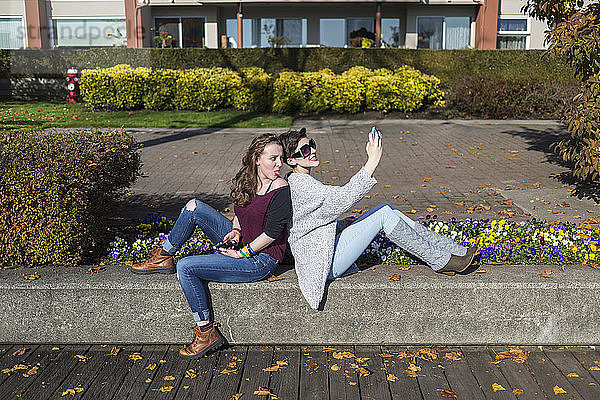 Zwei junge Frauen sitzen Rücken an Rücken und benutzen ihre Smartphones und machen Blödsinn; New Westminster  British Columbia  Kanada