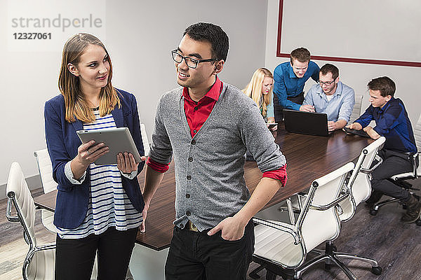 Zwei junge Geschäftsleute schauen auf ein Tablet  während sie in einem Konferenzraum mit Gleichaltrigen zusammenarbeiten; Sherwood Park  Alberta  Kanada