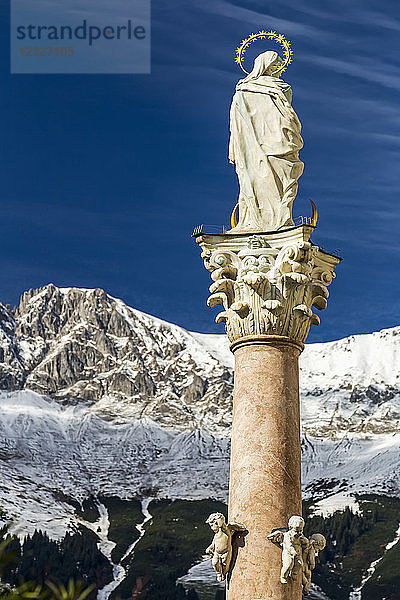 Steinstatue mit Heiligenschein auf der Spitze einer Säule mit schneebedecktem Gebirge und blauem Himmel im Hintergrund; Innsbruck  Tirol  Österreich