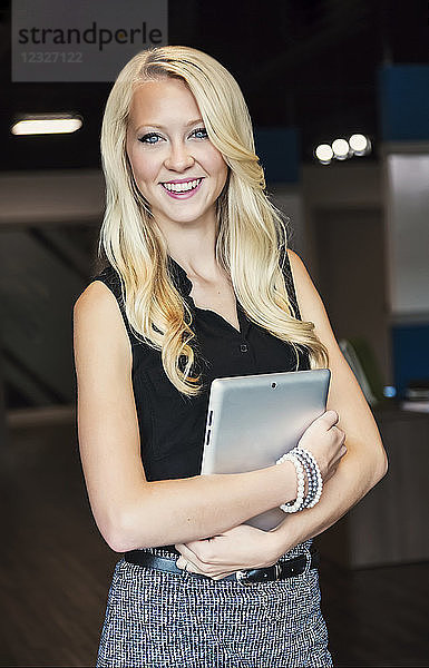 Business-Porträt einer schönen jungen Geschäftsfrau mit langen blonden Haaren  die ein Tablet hält und für die Kamera am Arbeitsplatz posiert; Sherwood Park  Alberta  Kanada