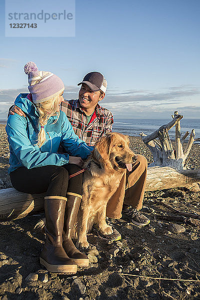 Ein junges Paar sitzt mit seinem Hund auf einem Stück Treibholz am Strand bei Sonnenuntergang; Anchorage  Alaska  Vereinigte Staaten von Amerika