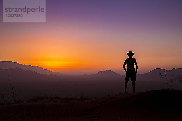 Silhouette eines Mannes mit Cowboyhut  der bei Sonnenuntergang auf die leuchtende Landschaft und den Himmel blickt; Sossusvlei  Hardap-Region  Namibia