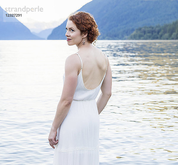 Eine Frau mit roten Haaren und einem weißen Kleid steht an einem Sommerabend im ruhigen Wasser des Alouette Lake; British Columbia  Kanada