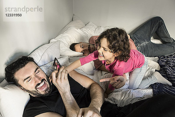 Daughter lying by man playing with father holding smart phone on bed in bedroom