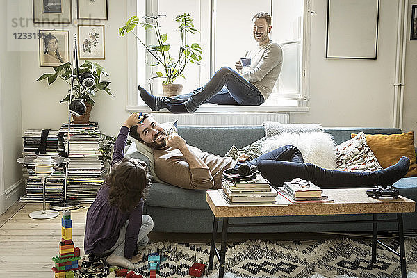 Girl playing with fathers while man sitting on window sill in living room