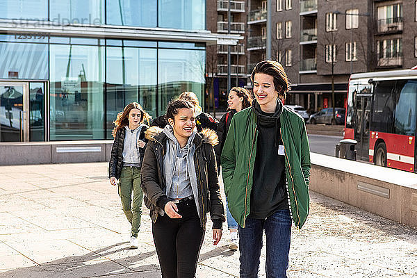 Smiling multi-ethnic teenagers wearing warm clothing walking on footpath in city during sunny day