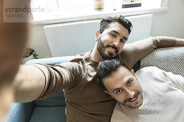Smiling gay couple taking selfie while relaxing on sofa in living room at home
