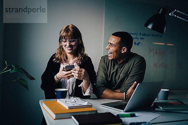 Cheerful multi-ethnic business colleagues sitting at desk with technologies at creative office