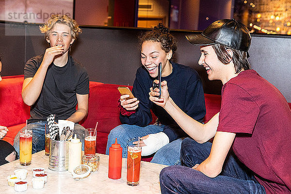 Cheerful teenage friends enjoying while using smart phones at restaurant