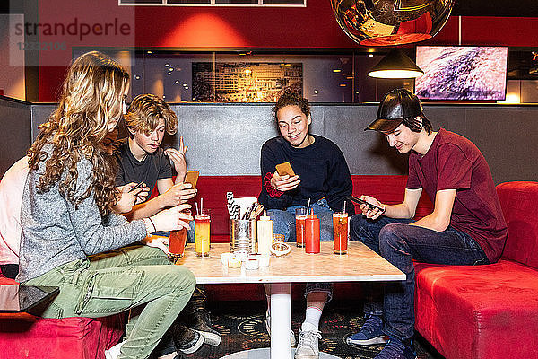 Multi-ethnic teenage friends using smart phone while sitting with mocktails on sofa at restaurant