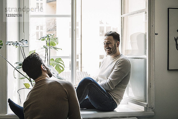 Happy gay man talking to boyfriend while sitting on windowsill at home