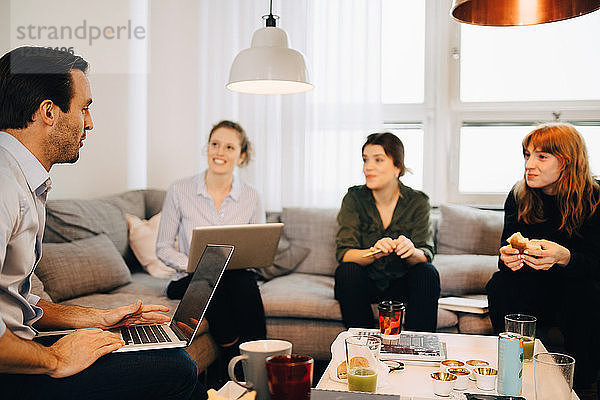 Female professionals looking at businessman sitting with laptop on sofa in creative office