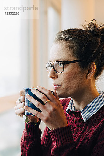 Thoughtful businesswoman drinking coffee while looking through window at creative office