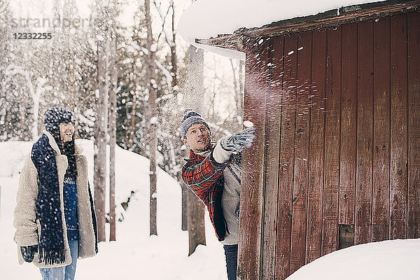 Friend looking at man throwing snowball while standing behind log cabin during winter