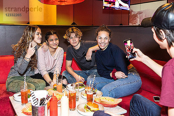 Teenage boy photographing multi-ethnic friends sitting on sofa at restaurant