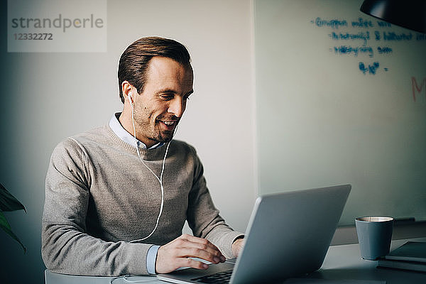 Smiling businessman using laptop while listening to in-ear headphones at creative office