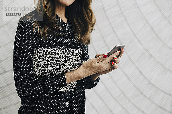 Midsection of businesswoman holding mobile phone while standing against wall