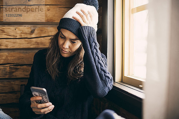 Mid adult woman using mobile phone while sitting by window in log cabin