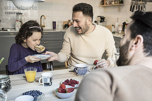 Laughing father holding plate while daughter eating pancake at table