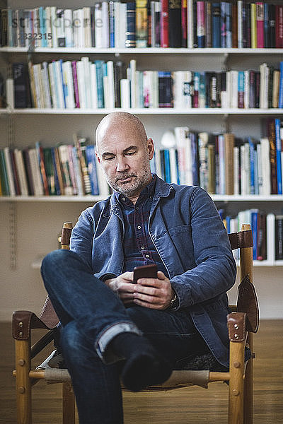Confident mature therapist using mobile phone while sitting against bookshelf at home office