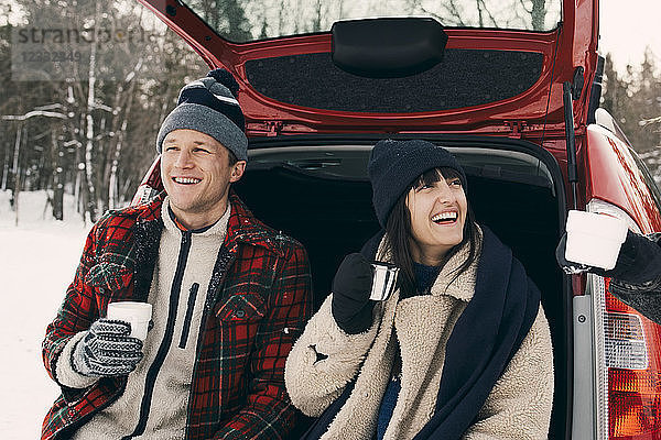 Happy woman having coffee while sitting with friends in car trunk during winter