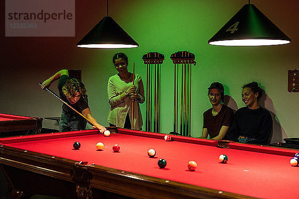 Smiling multi-ethnic friends looking at teenage boy aiming cue ball on illuminated red pool table