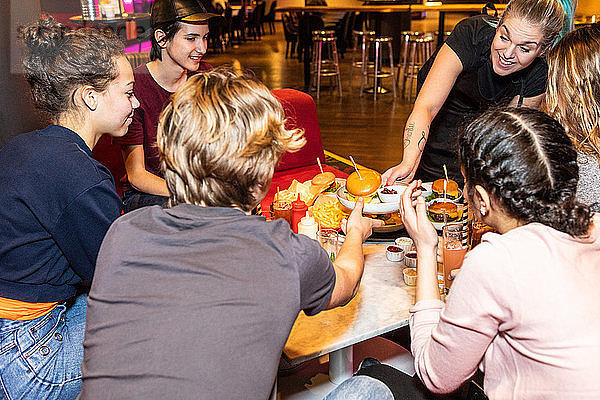 Smiling waitress serving food to multi-ethnic teenagers sitting at restaurant