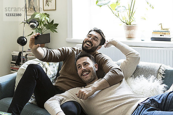 Happy gay couple taking selfie through mobile phone while relaxing on sofa at home