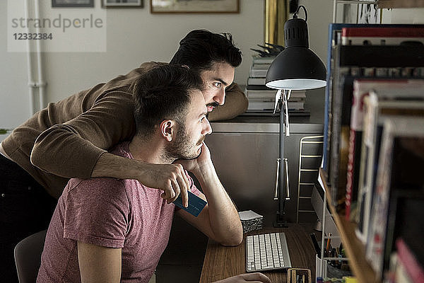 Gay couple shopping online through credit card on computer at illuminated desk in living room