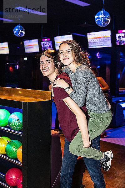 Portrait of smiling teenage boy giving piggyback ride to friend by rack at bowling alley