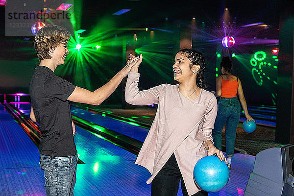 Smiling multi-ethnic friends giving high-five on illuminated parquet floor at bowling alley