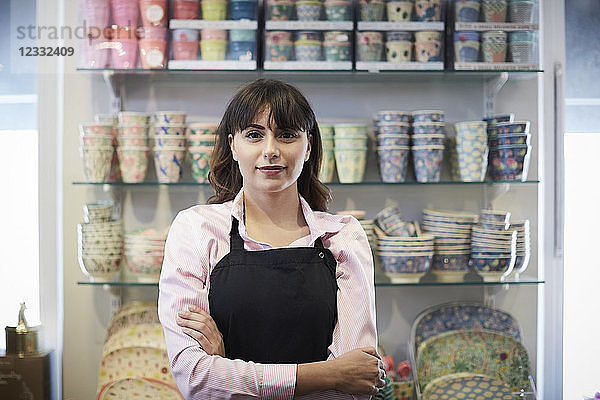 Portrait of confident saleswoman standing with arms crossed against crockery at boutique