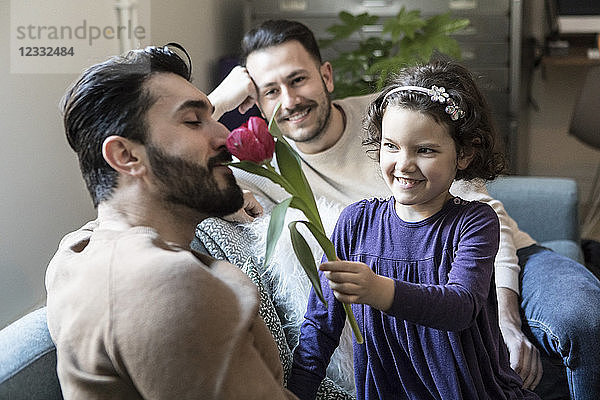 Smiling daughter making father smell tulip while sitting with man on sofa in living room