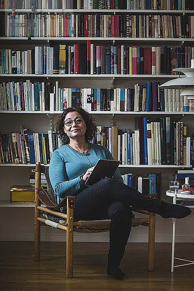 Portrait of female therapist holding digital tablet while sitting against bookshelf