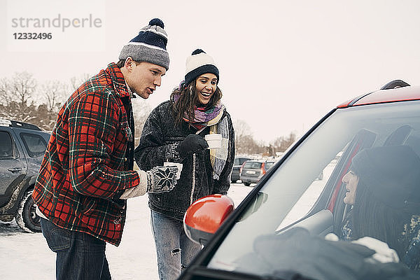 Happy man and woman having coffee while talking to friend sitting in car during winter