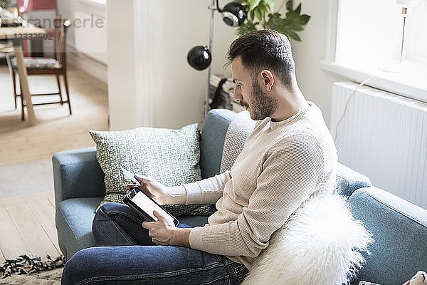 Young man doing online shopping on digital tablet through credit card while sitting on sofa at home