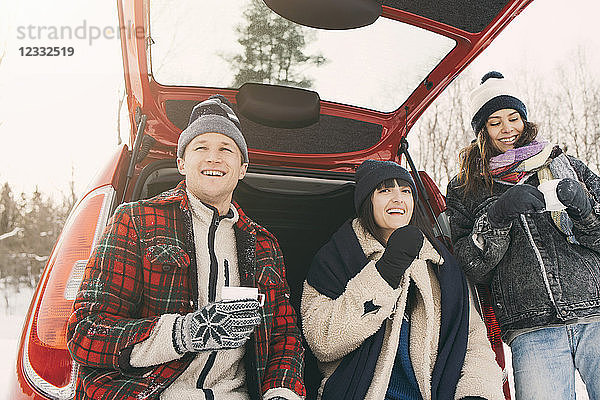 Low angle view of happy friends having coffee while resting in car trunk during winter