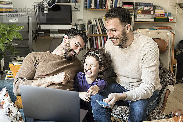 Happy fathers with daughter shopping online on laptop through credit card while sitting on chairs at home