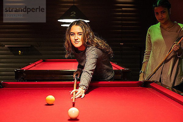 Smiling teenage girl aiming cue ball on illuminated red pool table by friend