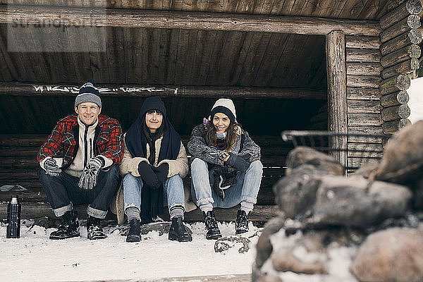 Full length portrait of friends sitting in log cabin against trees during winter