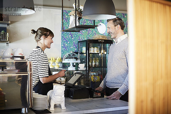 Smiling barista talking to customer while using cash register at checkout counter in cafe