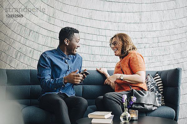 Happy male and female colleagues talking while sitting on couch against wall