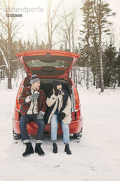 Full length of friends having coffee while sitting in car trunk on snowy field