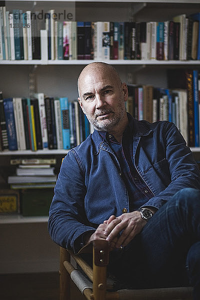 Portrait of confident mature therapist sitting against bookshelf at home office