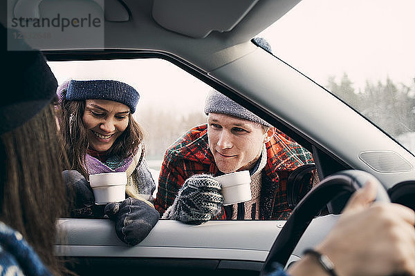 Smiling man and woman talking to friend sitting in car while having coffee during winter